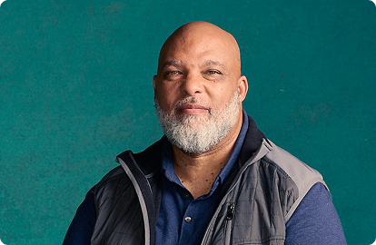 A black man with a beard and blue shirt smiles confidently at the camera.